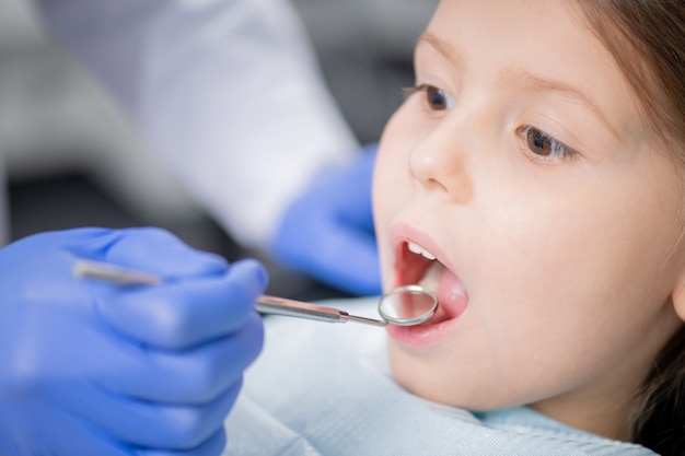 Mano enguantada del dentista profesional con espejo dental mientras examina los dientes de la niña con la boca abierta