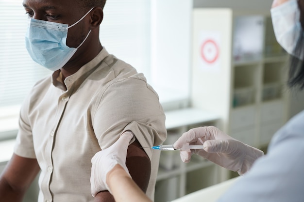 Mano enguantada de enfermera en uniforme y máscara protectora que vacuna al chico africano sentado frente a ella en el consultorio médico de las clínicas contemporáneas