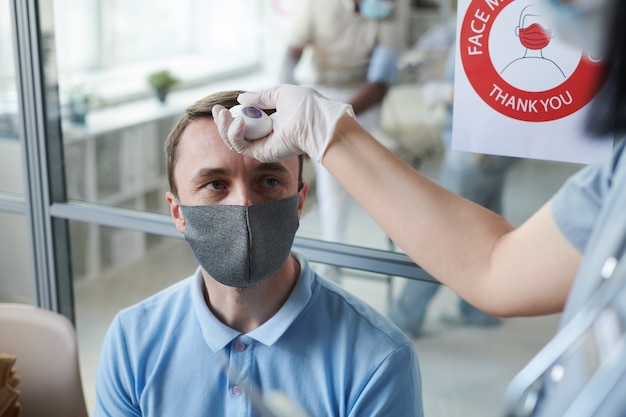 Mano enguantada de la joven doctora o enfermera en uniforme midiendo la temperatura corporal del paciente masculino en máscara protectora y ropa informal