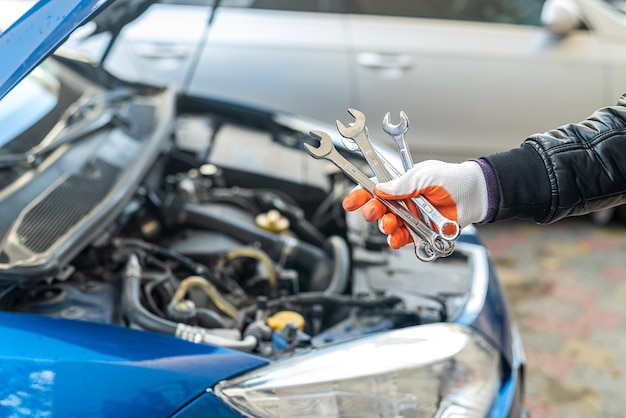 La mano enguantada del mecánico del hombre sostiene las herramientas del automóvil cerca de un automóvil pintado con el capó abierto. Concepto de herramientas. Concepto de reparación de automóviles