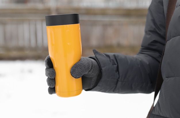 Una mano enguantada sostiene un termo amarillo con una bebida caliente