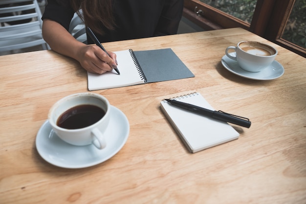 Una mano está escribiendo en un cuaderno con tazas de café en la mesa de madera