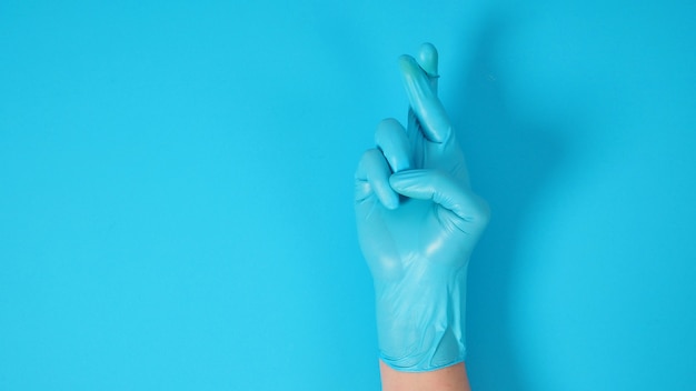 Mano está haciendo el signo de la mano con los dedos cruzados y usando guantes quirúrgicos sobre fondo azul.