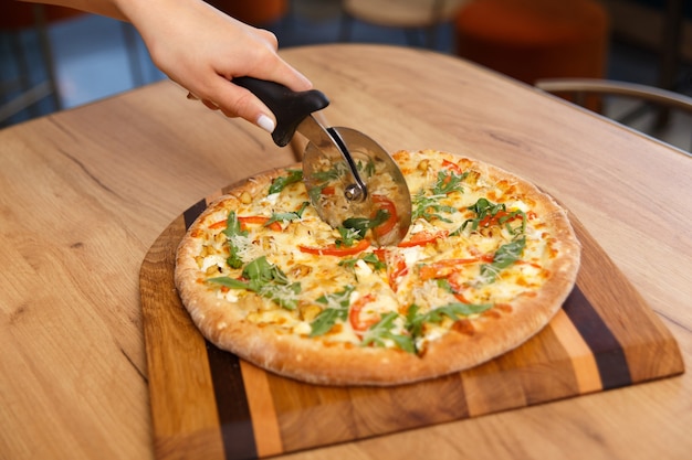 Mano femenina con cuchillo circular cortando rebanadas de pizza recién sobre tabla de madera en la cocina, enfoque suave. Mano rebanar pizza con cortador en la mesa.