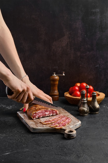 Mano femenina con cuchillo rebanar salchicha de cerdo ahumada en tripa orgánica sobre tabla de cortar de madera