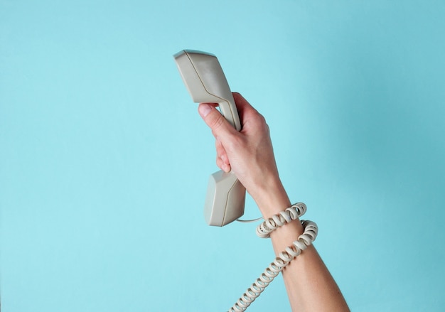 Mano femenina envuelta en cable sostiene el tubo del teléfono contra un azul