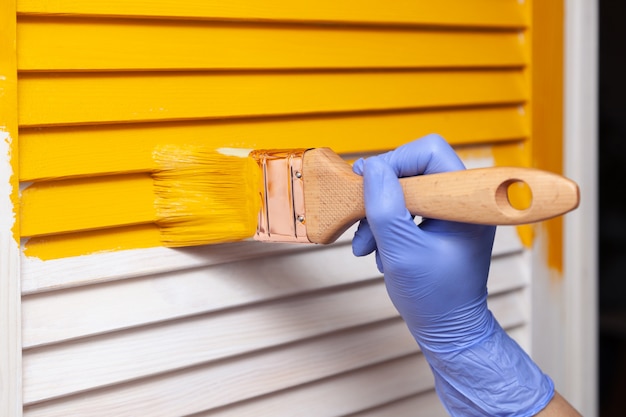 Mano femenina en guante de goma con pincel pintando puerta de madera con pintura amarilla