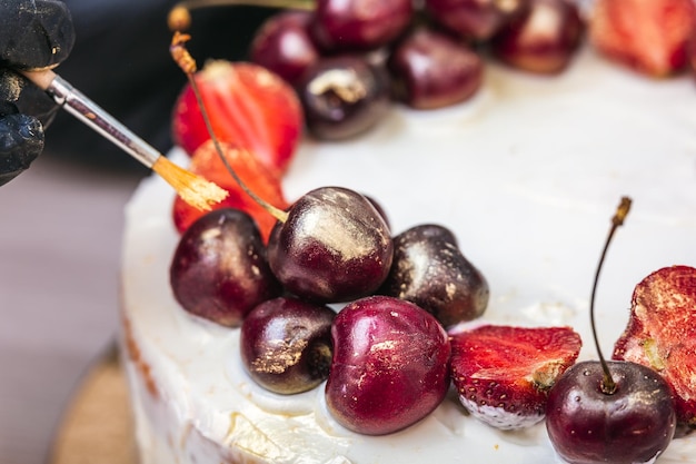 Mano femenina en guante negro pone oro comestible en cerezas y fresas encima de tarta de queso con c