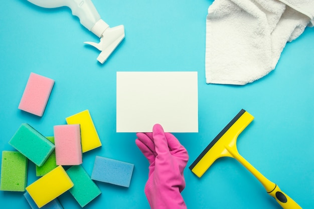 La mano femenina en guantes de color rosa tiene una tarjeta vacía y guantes, un spray, esponjas, un raspador para ventanas, una toalla sobre un fondo azul. Concepto de servicio de limpieza. Copia espacio Endecha plana, vista superior