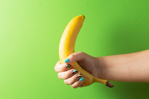 Mano femenina con manicura colorida con plátano sobre fondo verde