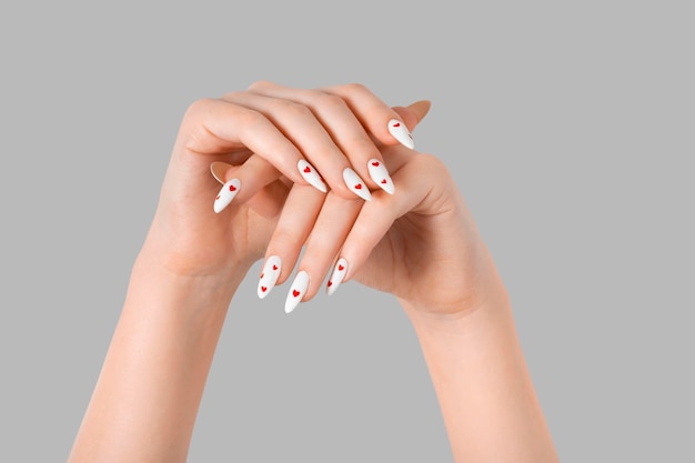 Mano femenina de manicura con esmalte de gel, uñas largas blancas y diseño de corazones rojos sobre un fondo gris