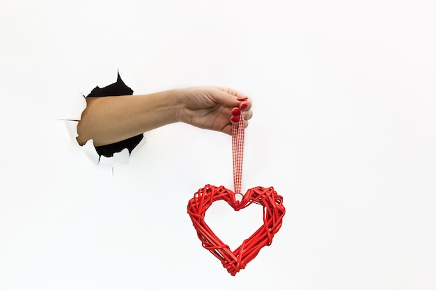 Una mano femenina con una manicura roja a través de papel blanco rasgado sostiene un corazón rojo. Mano a través de papel blanco rasgado