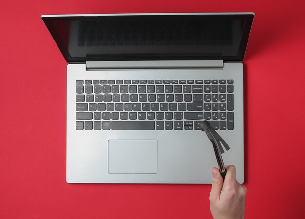 Mano femenina con un martillo golpea un teclado de computadora portátil sobre un fondo rojo.