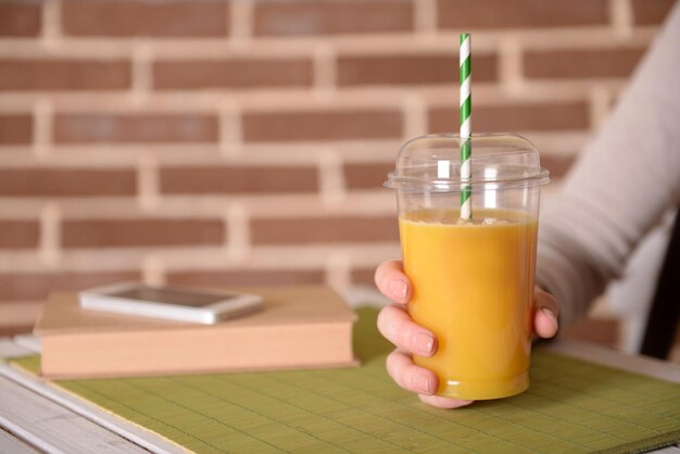 Mano femenina en la mesa con comida rápida taza cerrada de jugo de naranja sobre fondo de pared de ladrillo