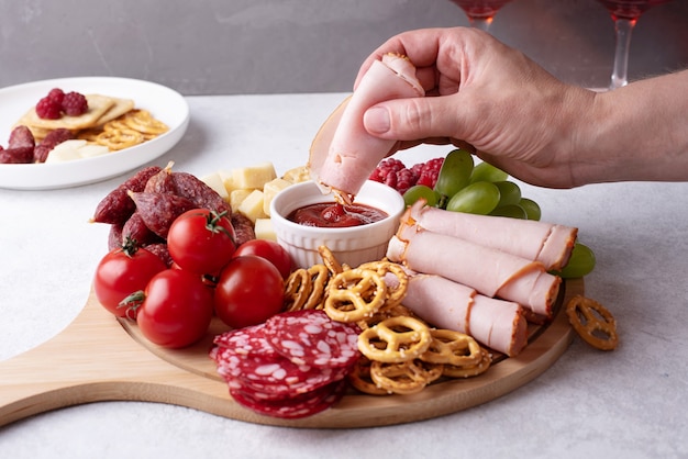 Mano femenina mojar una rebanada de jamón en salsa sobre placa redonda de charcutería con salchicha, queso, galletas y fruta, platos con aperitivo, primer plano.