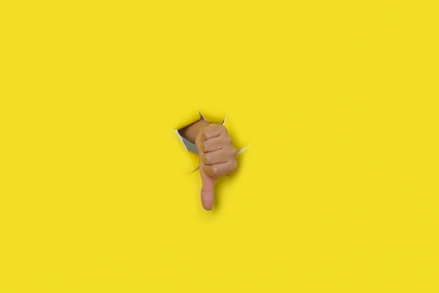 Mano femenina con el pulgar hacia abajo como señal de desaprobación saliendo del agujero de un papel amarillo rasgado