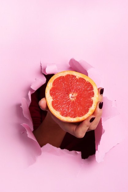 Mano femenina que lleva a cabo la mitad del pomelo a través del fondo de papel rosado rasgado. Jugo de naranja fresco.