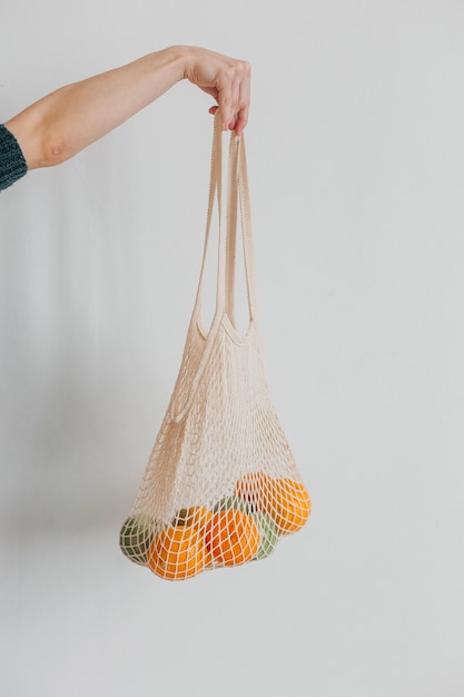 Mano femenina que sostiene la bolsa ecológica de malla con frutas en la pared blanca Concepto de compras ecológicas.