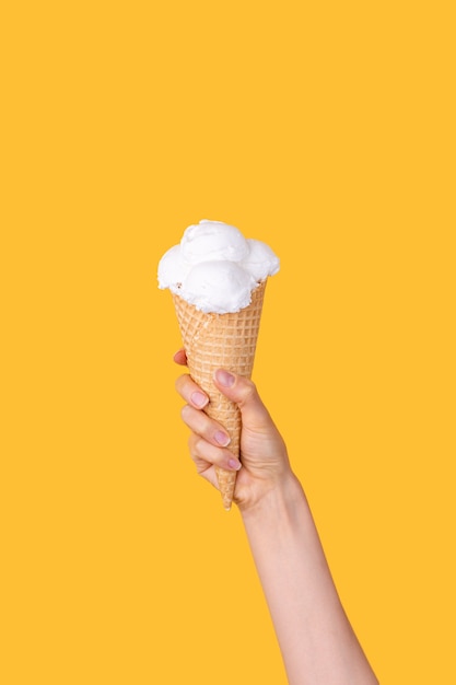 Mano femenina que sostiene el cono de galleta con cucharas de helado de vainilla como antorcha sobre fondo amarillo brillante