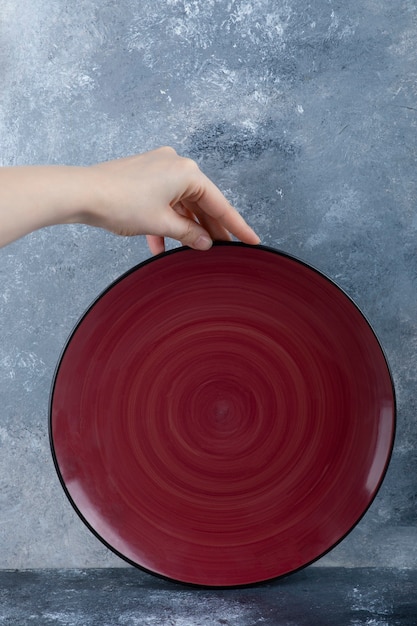 Mano femenina que sostiene el plato vacío rojo en mármol.