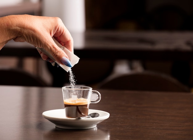 Mano femenina con sobres de azúcar vertidos en la taza de café, en la parte superior de la mesa de madera