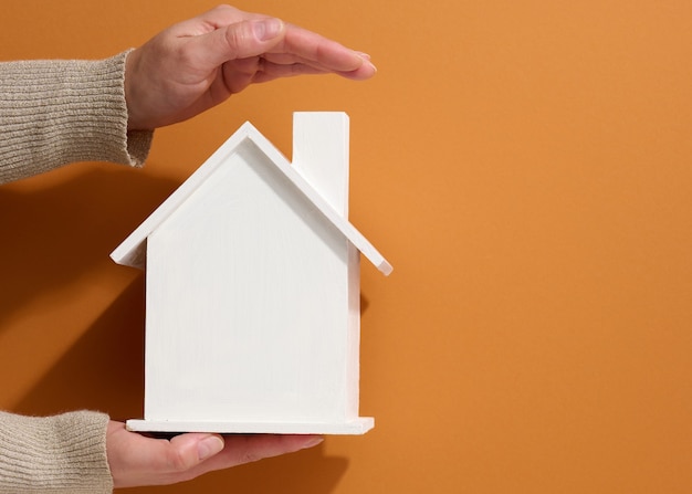 Mano femenina sostenga una casa de madera en miniatura blanca sobre un fondo marrón. Concepto de seguro inmobiliario, protección del medio ambiente, felicidad familiar.