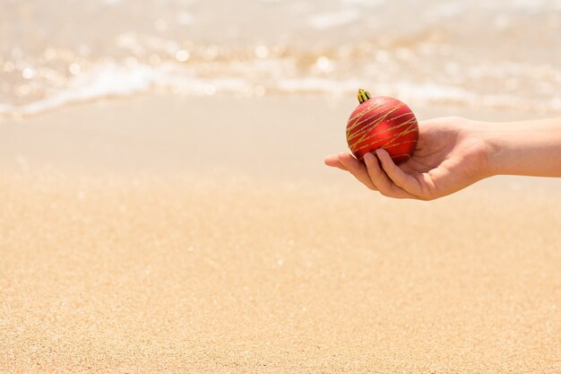 Mano femenina sosteniendo una bola roja de Navidad en el fondo del mar, vacaciones de Navidad en la playa