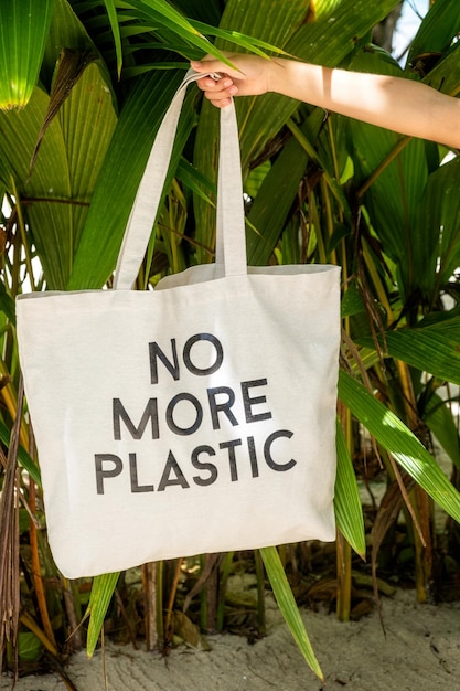 Mano femenina sosteniendo una bolsa ecológica con un letrero sin más plástico en un fondo de follaje tropical verde