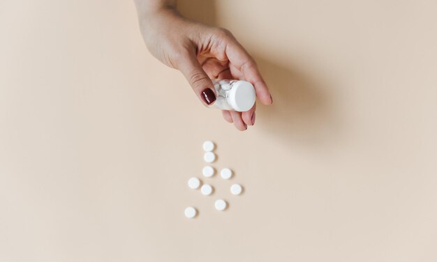 Mano femenina sosteniendo una botella de pastillas. fondo beige. Frasco de vidrio para medicamentos con pastillas blancas