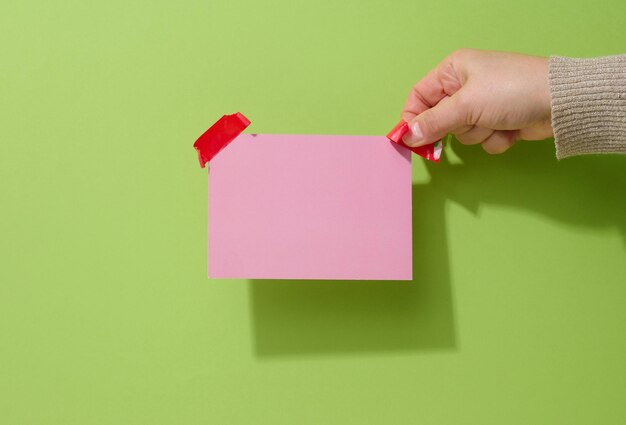 Mano femenina sosteniendo una hoja de papel rosa en blanco con cinta adhesiva roja sobre fondo verde