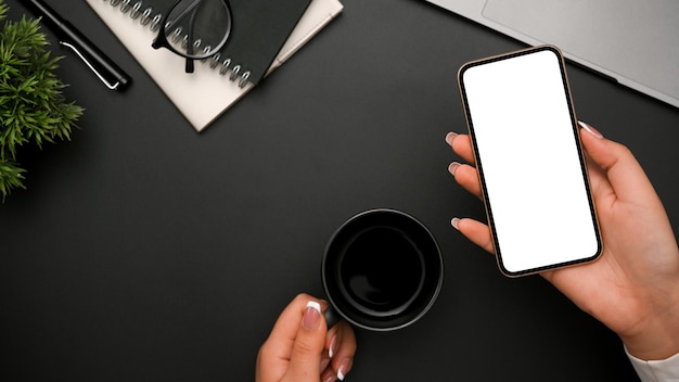 Mano femenina sosteniendo una maqueta de pantalla blanca de teléfono inteligente y una taza de café sobre un escritorio de oficina negro moderno