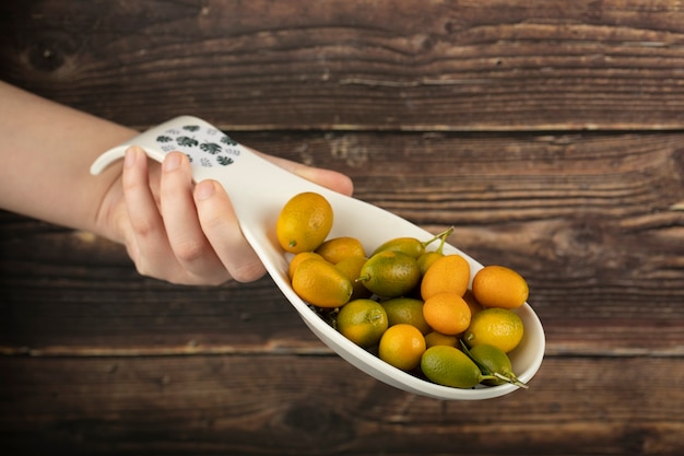 Foto mano femenina sosteniendo un plato pequeño de kumquats frescos en la superficie de madera.