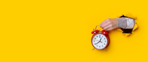 Mano femenina sosteniendo un reloj rojo en un agujero sobre un fondo amarillo. Bandera.