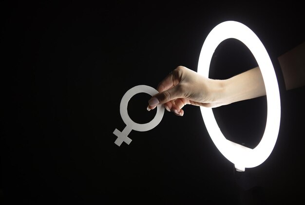 Mano femenina sosteniendo el símbolo de Venus a través de la lámpara de anillo de led en fondo negro Feminismo
