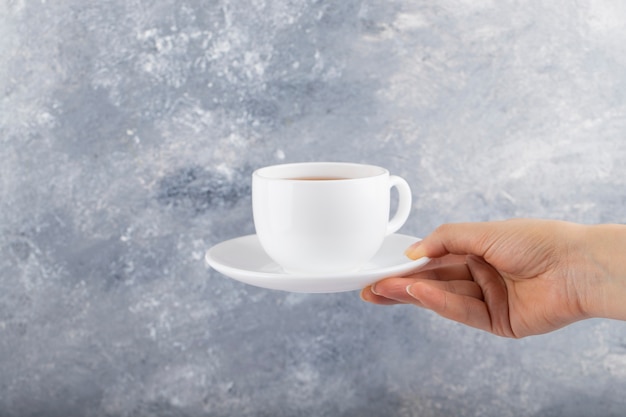 Mano femenina sosteniendo una taza blanca de té negro sobre una mesa de piedra.