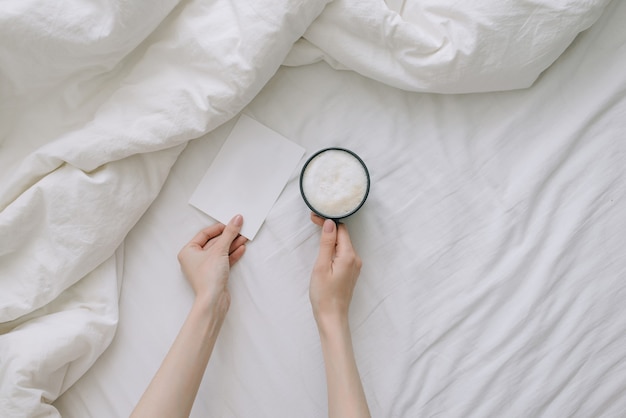 Mano femenina sosteniendo una taza de café y una postal en ropa de cama blanca. Vista superior.