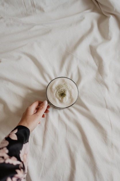 Foto una mano femenina sosteniendo una taza de matcha latte con una espuma blanca en la parte superior de la cama blanca