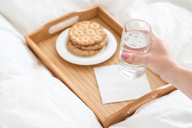 Mano femenina sosteniendo un vaso de agua sobre la bandeja con galletas en la cama