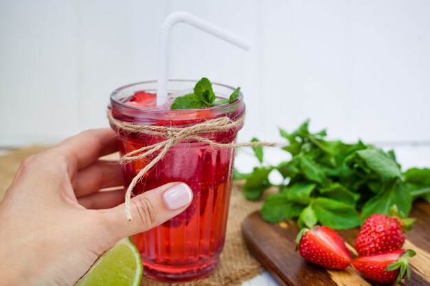 Mano femenina sostiene bebida de fresa en un vaso con limón y menta