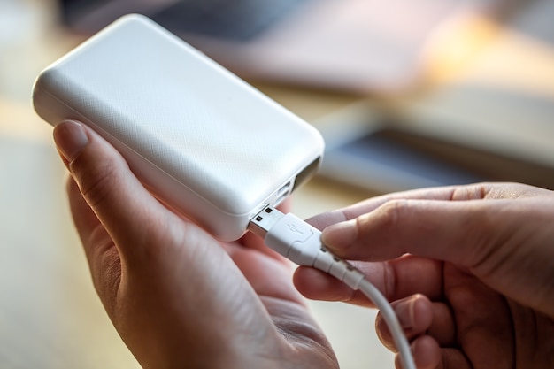 Una mano femenina sostiene un cable USB blanco y un banco de energía blanco, cargando el teléfono con un banco de energía.