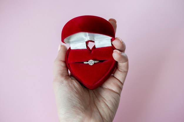 Mano femenina sostiene caja en forma de corazón rojo con anillo de compromiso sobre fondo rosa