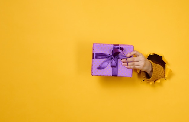 Mano femenina sostiene una caja con un regalo sobre un fondo amarillo parte del cuerpo sobresale de un agujero rasgado en un fondo de papel Felicitaciones vacaciones sorpresax9