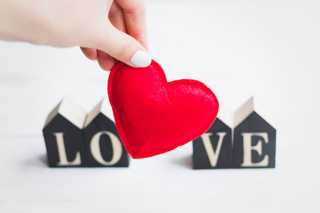 La mano femenina sostiene un corazón rojo en el fondo de la palabra amor. Tarjeta de San Valentín sobre fondo blanco de madera