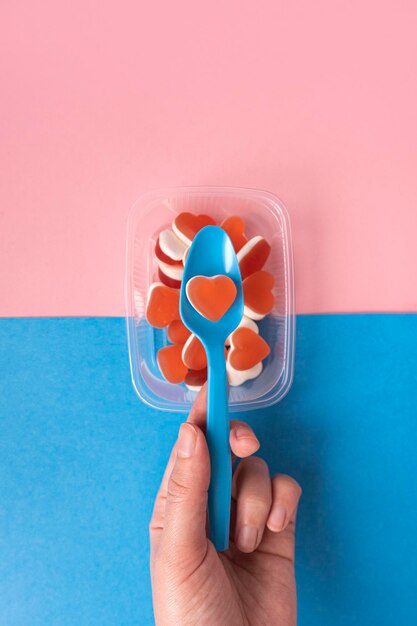 La mano femenina sostiene una cuchara azul de plástico con mermelada de gelatina en forma de corazón rojo Concepto de comida rápida