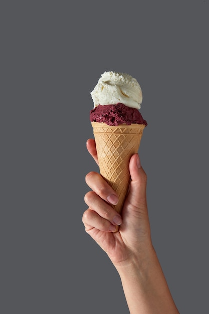 Una mano femenina sostiene helado de frutos rojos y helado de leche blanca en un cono de galleta sobre una pared gris. Concepto de verano con espacio de copia.