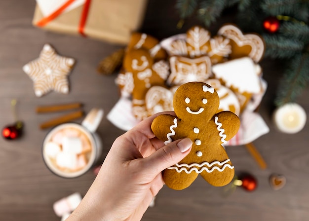 Una mano femenina sostiene a un hombre de pan de jengibre sobre un fondo de una canasta de pan de jengibre una taza de cacao con malvaviscos una caja de regalo y ramas de árboles de Navidad