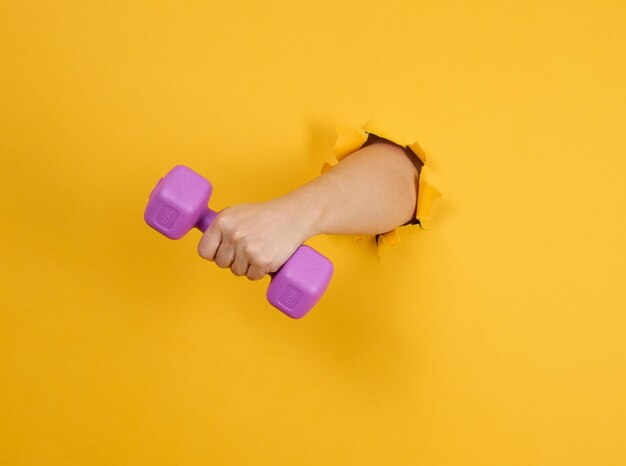 mano femenina sostiene una manivela de plástico de un kilogramo en un fondo amarillo una parte del cuerpo sobresale de un agujero rasgado en un fondo de papel estilo de vida saludable deportes