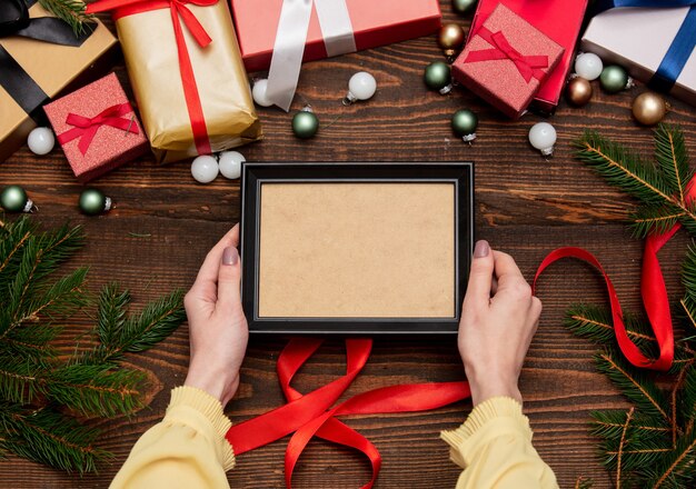 Mano femenina sostiene el marco de imagen cerca con regalos de navidad y ramas de pino con adornos en la mesa de madera