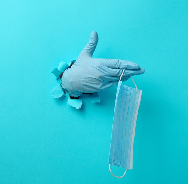 Una mano femenina sostiene una máscara médica desechable para protegerse contra los virus durante una epidemia y una pandemia. Parte del cuerpo sobresale de un agujero rasgado en papel azul