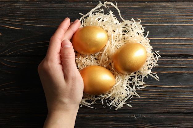 Mano femenina sostiene nido con huevos de oro en madera, vista superior